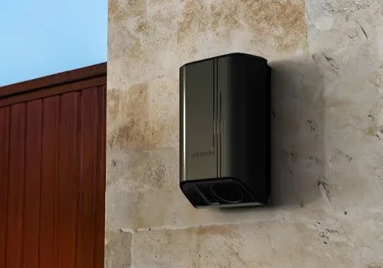 Modern black wall-mounted mailbox on a stone wall beside a wooden gate.